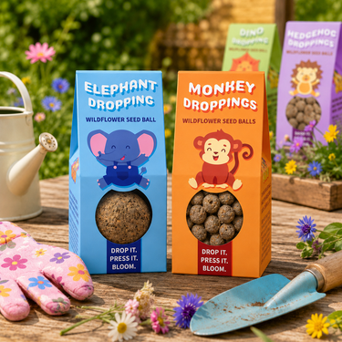 Two boxes of wildflower seed balls displayed on a wooden table in a garden setting, with gardening gloves, a watering can, and a small trowel around them. The boxes are blue and orange with cartoon animal illustrations.