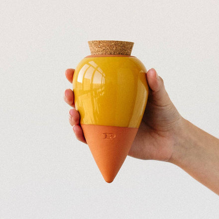 Hand holding a yellow and orange ceramic container with a cork lid on a light gray background