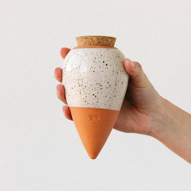Hand holding a ceramic jar with a speckled white body and terracotta base, against a plain background.