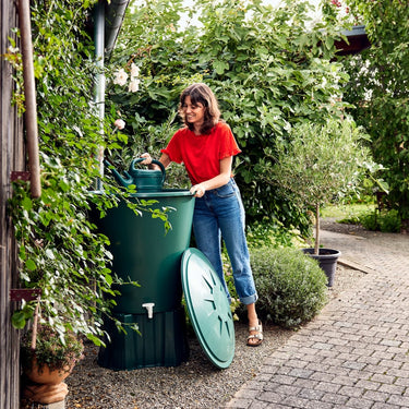 Woman in a red shirt and blue jeans interacting with a green compost bin in a garden setting.