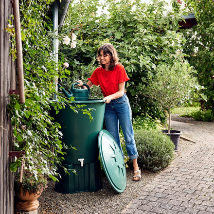 Woman in a red shirt and blue jeans interacting with a green compost bin in a garden setting.