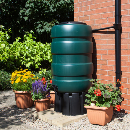 Green rain barrel attached to a brick wall with potted plants around