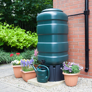 Green rainwater harvesting tank attached to a brick wall with potted plants nearby.