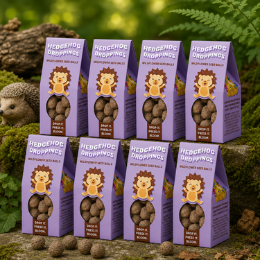 Eight purple bags of Hedgehog Droppings wildflower seed balls arranged on a mossy log outdoors, with a hedgehog visible on the left and loose seed balls in the foreground.